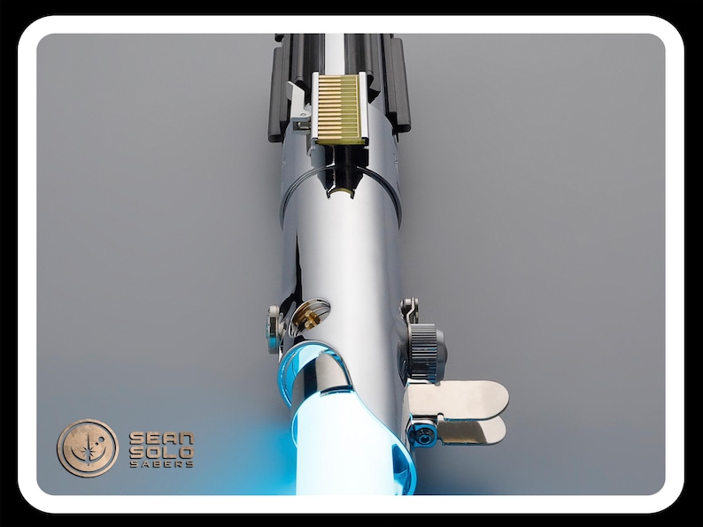 Puede incluir: Un sable de luz cromado con una hoja azul brillante. El mango presenta detalles intrincados, incluyendo una secci&oacute;n dorada y varios componentes met&aacute;licos. El logotipo "Sean Solo Sabers" es visible en la esquina inferior izquierda.
