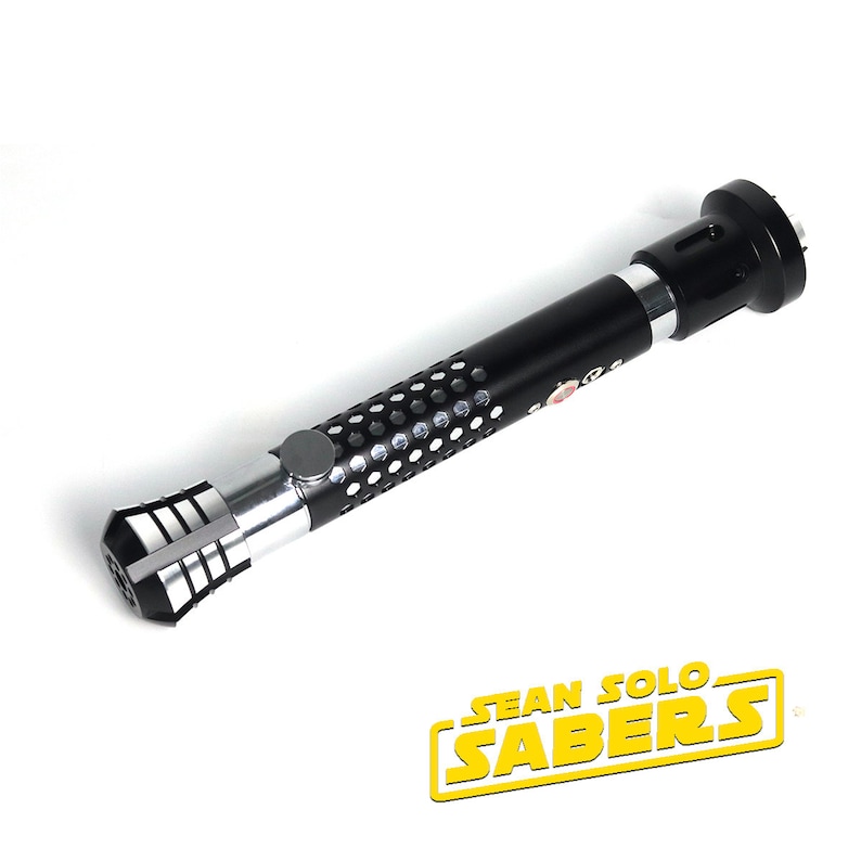 May include: A black lightsaber hilt with a silver and black emitter and a silver and black pommel. The hilt has a textured, honeycomb-like design. The hilt is labeled "SEAN SOLO SABERS".