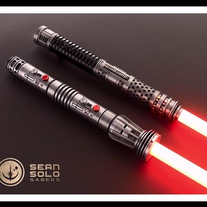 May include: Two metallic lightsaber hilts with glowing red blades. The hilts feature intricate silver and black detailing, circular vents, and red activation buttons. A logo in the corner reads Sean Solo Sabers with a circular star emblem.