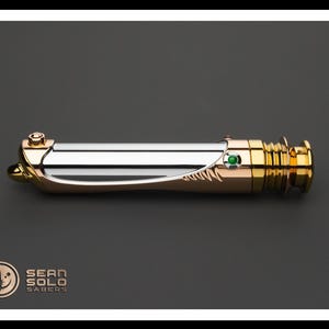 May include: A metallic lightsaber with gold, silver, and copper accents. The hilt features a green gem and intricate detailing. The product is displayed on a dark gray background with the "Sean Solo Sabers" logo.