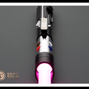 Op de afbeelding: Een zwart lightsaber met een roze blade. De lightsaber heeft een zilveren handvat met een zwarte grip en een zilveren emitter. De lightsaber is aan en de blade gloeit roze.