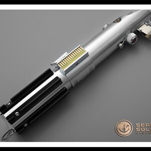 Puede incluir: Un sable de luz plateado con empu&ntilde;aduras negras y un detalle dorado. El accesorio tiene un acabado met&aacute;lico y est&aacute; sobre un fondo gris. El logotipo de Sean Solo Sabers est&aacute; en la esquina inferior derecha.