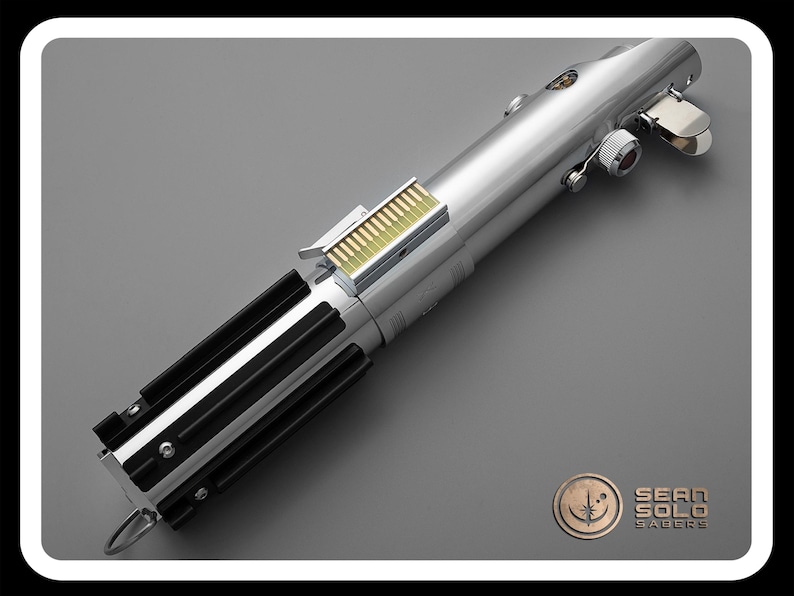 Puede incluir: Un sable de luz plateado con empu&ntilde;aduras negras y un detalle dorado. El accesorio tiene un acabado met&aacute;lico y est&aacute; sobre un fondo gris. El logotipo de Sean Solo Sabers est&aacute; en la esquina inferior derecha.