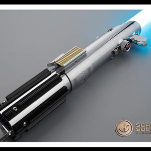 Puede incluir: Un detallado sable de luz de r&eacute;plica con un acabado plateado pulido y detalles en negro. El mango presenta intrincados detalles mec&aacute;nicos, y una hoja azul brillante se extiende desde el emisor. El logotipo de Sean Solo Sabers es visible.