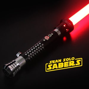 May include: A black lightsaber with a red blade and a silver handle. The lightsaber has a textured grip and a small button on the side. The lightsaber is lit up and the blade is glowing red. The lightsaber is on a black surface. The text "SEAN SOLO SABERS" is in yellow letters on the surface.