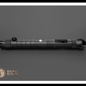 May include: A black lightsaber with a silver button and a black handle. The lightsaber is on a gray surface.