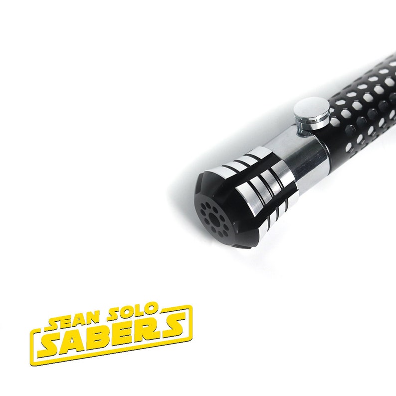 May include: Close-up of the black and silver hilt of a lightsaber with a textured black handle. The hilt has a silver band with a series of grooves and a black cap with a circular pattern of holes. The text "SEAN SOLO SABERS" is visible in the lower left corner of the image.