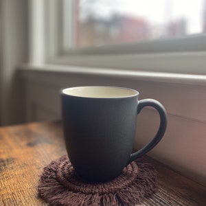 Pode incluir: Uma caneca de cer&acirc;mica cinza escura com interior branco est&aacute; sobre um descanso de copo de croch&ecirc; marrom com franjas.