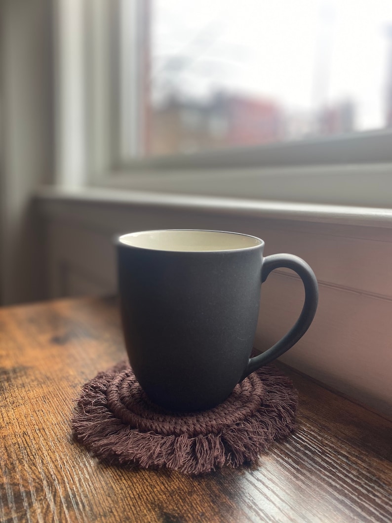 Pode incluir: Uma caneca de cer&acirc;mica cinza escura com interior branco est&aacute; sobre um descanso de copo de croch&ecirc; marrom com franjas.