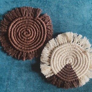 Pode incluir: Dois tapetes de croch&ecirc; feitos &agrave; m&atilde;o. Um &eacute; castanho com uma borda de franja. O outro &eacute; branco com um design de semic&iacute;rculo castanho e uma borda de franja.