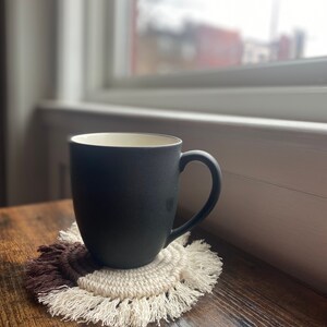 Pode incluir: Uma caneca de cer&acirc;mica preta com interior branco est&aacute; sobre um descanso de copo de croch&ecirc; marrom e branco com franjas. A caneca est&aacute; sobre uma superf&iacute;cie de madeira.