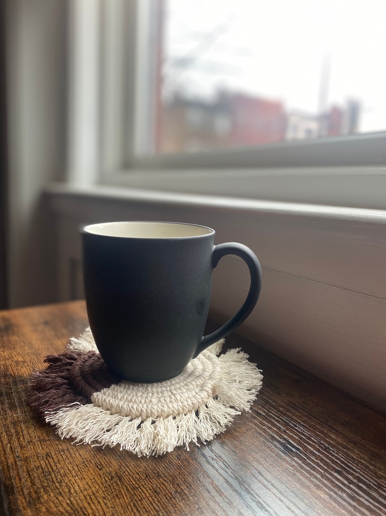 Pode incluir: Uma caneca de cer&acirc;mica preta com interior branco est&aacute; sobre um descanso de copo de croch&ecirc; marrom e branco com franjas. A caneca est&aacute; sobre uma superf&iacute;cie de madeira.