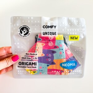 Può includere: Una mascherina facciale origami riutilizzabile in una confezione di plastica trasparente. La mascherina presenta un colorato motivo a gatti. La confezione include le parole "COMFY UNIQUE" e "NEW". La mascherina è etichettata "Non Medical" e "Adult Size".