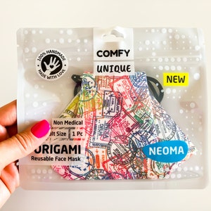 Può includere: Una mascherina riutilizzabile in una confezione di plastica trasparente. La mascherina presenta un design colorato di timbri di viaggio. La confezione riporta le parole "COMFY UNIQUE" e "NEW". La mascherina è etichettata "ORIGAMI" e "NEOMA".