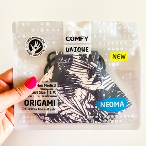 Può includere: Una mascherina origami riutilizzabile in una confezione di plastica trasparente. La mascherina ha un motivo a foglie tropicali in bianco e nero. La confezione include il testo "COMFY UNIQUE" e "NEW".