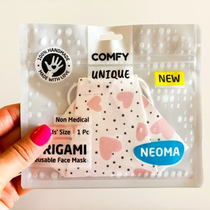Può includere: Una mascherina origami bianca e rosa a forma di cuore in una confezione di plastica trasparente. La confezione include le parole "COMFY", "UNIQUE" e "NEW". La mascherina è etichettata come "Non Medical" e "Reusable Face Mask". Il marchio "NEOMA" è visibile.