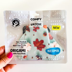 Può includere: Una mascherina per il viso in una confezione di plastica trasparente. La mascherina ha uno sfondo azzurro chiaro con un motivo di foglie e bacche rosse. La confezione riporta le scritte "COMFY UNIQUE" e "NEW". La mascherina è etichettata "ORIGAMI" e "NEOMA".