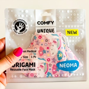 Può includere: Una mascherina facciale riutilizzabile origami a tema unicorno rosa in una confezione di plastica trasparente. La confezione riporta le parole "COMFY", "UNIQUE" e "NEW". La mascherina è decorata con unicorni, arcobaleni e stelle.