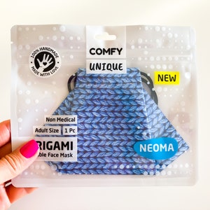 Può includere: Una mascherina origami blu e bianca in una confezione di plastica trasparente. La mascherina ha un motivo a maglia ed è etichettata "COMFY UNIQUE" e "NEW". La confezione include anche il testo "100% HANDMADE MADE WITH LOVE". Il marchio "NEOMA" è visibile.