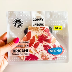Può includere: Una maschera facciale origami riutilizzabile in una confezione di plastica trasparente. La maschera presenta un motivo floreale nei toni del rosso, arancione e beige. La confezione riporta le scritte "COMFY UNIQUE" e "NEW". Il marchio "NEOMA" è visibile.