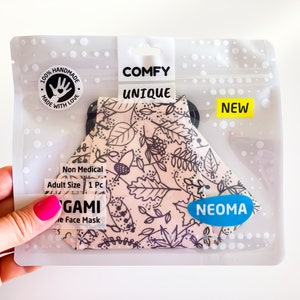 Può includere: Una mascherina per il viso in una confezione di plastica trasparente. La mascherina ha uno sfondo beige con un motivo a foglie nere. La confezione riporta "COMFY UNIQUE" e "NEW". La mascherina è etichettata "Non Medical" e "Adult Size 1 Pc".