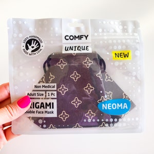 Può includere: Una mascherina origami riutilizzabile in una confezione di plastica trasparente. La mascherina è viola scuro con un motivo floreale dorato. La confezione riporta le parole "COMFY UNIQUE" e "NEW". La mascherina è etichettata come "Non Medical" e "Adult Size".