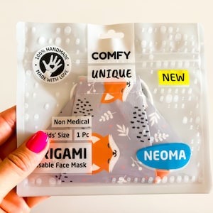 Può includere: Una mascherina per il viso in una confezione di plastica trasparente. La mascherina ha uno sfondo grigio con volpi arancioni e disegni floreali bianchi. La confezione riporta le scritte "COMFY UNIQUE" e "NEW". La mascherina è etichettata "Non Medical" e "Kids' Size".