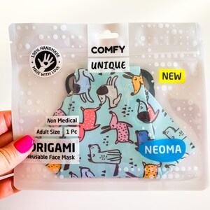 Può includere: Una mascherina origami riutilizzabile in una confezione di plastica trasparente. La mascherina ha uno sfondo azzurro con un motivo di cani a cartoni animati colorati. La confezione riporta le scritte "COMFY UNIQUE" e "NEW".