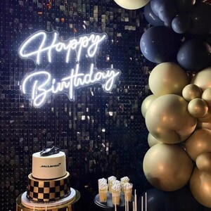 May include: A birthday cake with a black and gold checkered base and a white top, topped with a black race car. A neon sign displays "Happy Birthday". Gold, black, and blue balloons decorate the background. The cake is on a gold stand.