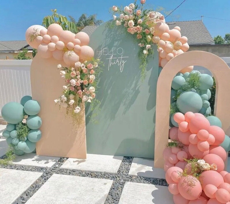 May include: A backdrop for a party with two arches, one peach and one light green. The peach arch is decorated with a cluster of pastel pink and white balloons. The green arch is decorated with a cluster of light green and pink balloons. The text "Hello Thirty" is written on the green arch.