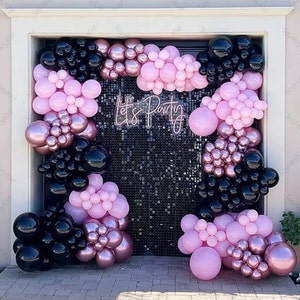 May include: A party backdrop with black, pink, and rose gold balloons. The balloons are arranged in clusters and arches around a black sequin wall. The words "Let's Party" are displayed in a neon sign.