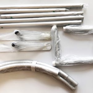 May include: A set of silver metal poles, curved poles, and base plates for a pop-up backdrop stand. The poles are packaged in plastic bags.