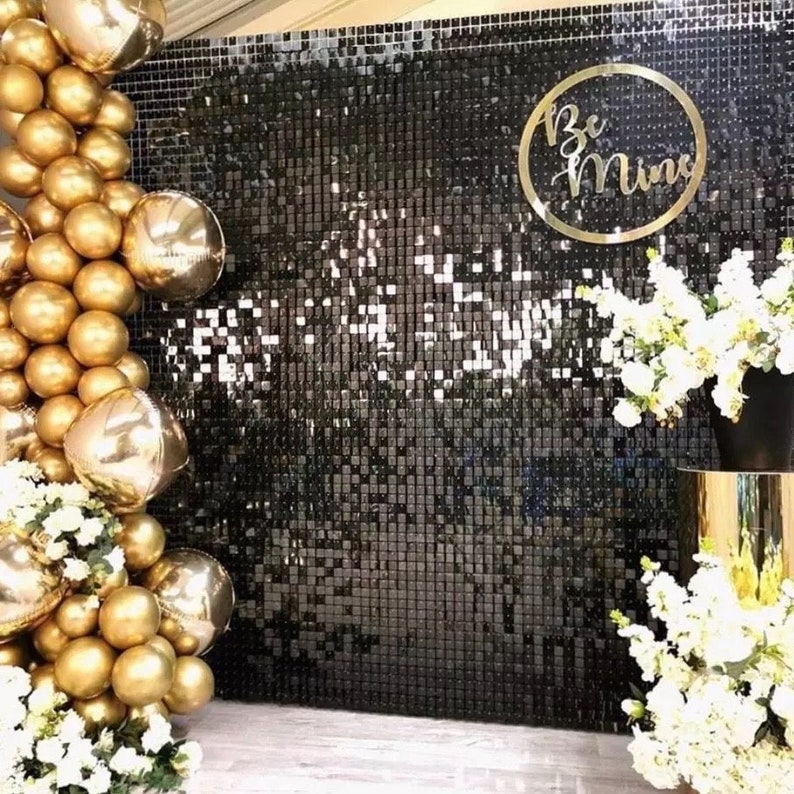 May include: A black sequin wall backdrop with gold balloons and white flowers. A gold circular sign with the words "Be Mine" is on the wall. The balloons are a mix of round and star-shaped.