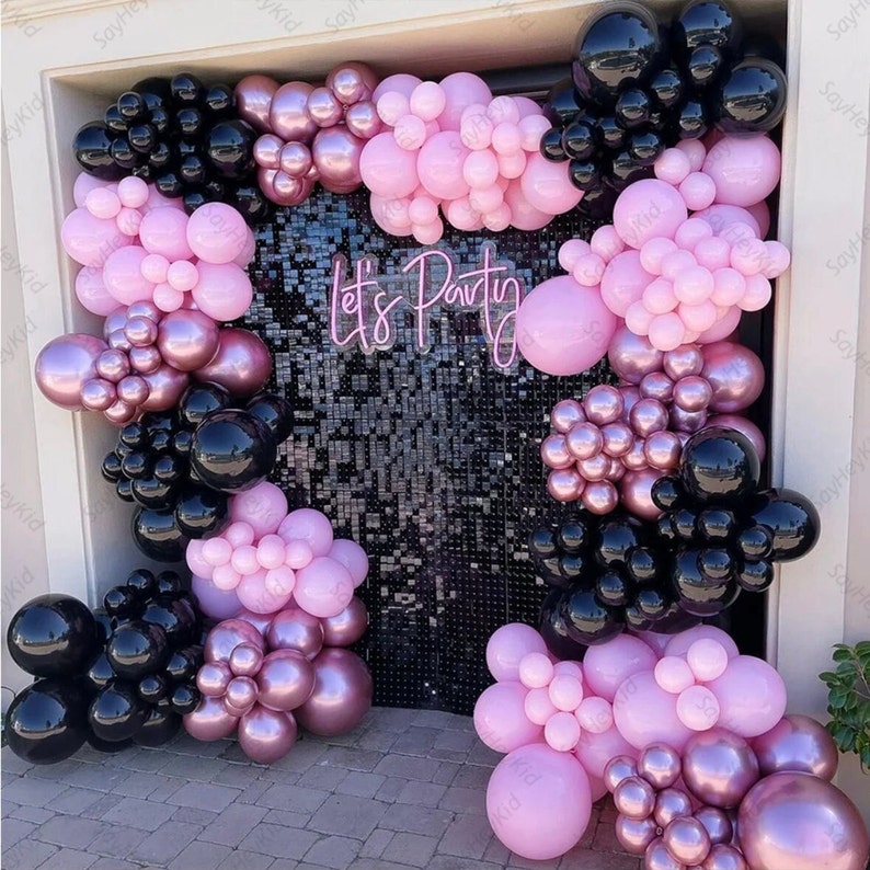 May include: A party backdrop with a black sequin wall, decorated with pink, black, and rose gold balloon clusters. The words "Let's Party" are in pink neon. The balloons are arranged in arches and clusters, creating a festive atmosphere.