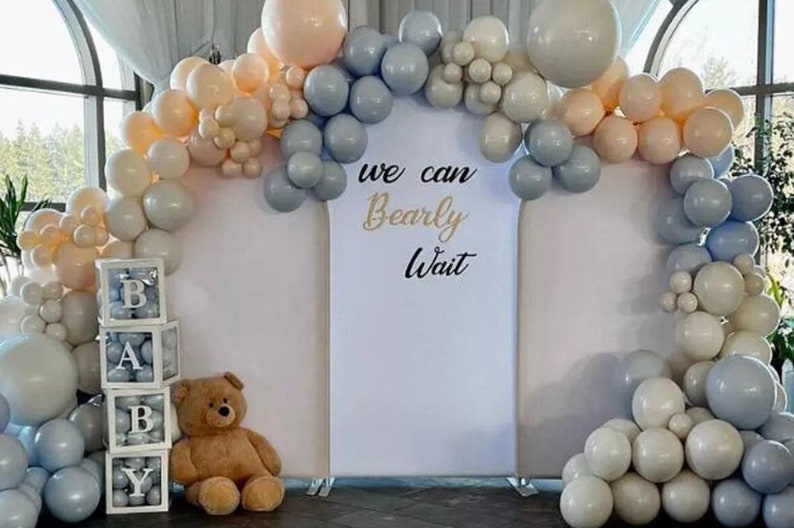 May include: A white backdrop with the text "We can bearly wait" decorated with a balloon arch of blue, white, and beige balloons.  A teddy bear and a stack of clear boxes with the letters "BABY" are in front of the backdrop.