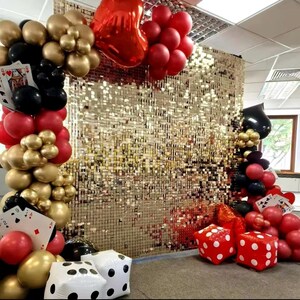 K&ouml;nnte beinhalten: Ein goldfarbener Sequin-Hintergrund mit roten, schwarzen und goldenen Ballons. Es gibt auch gro&szlig;e rote W&uuml;rfel und Spielkarten. Die Ballons sind in einer Bogenform angeordnet.