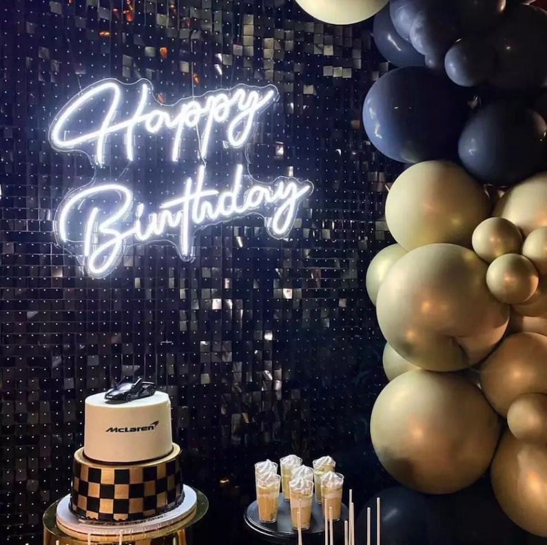 May include: A birthday cake with a black and gold checkered base and a white top, topped with a black race car. A neon sign displays "Happy Birthday". Gold, black, and blue balloons decorate the background. The cake is on a gold stand.