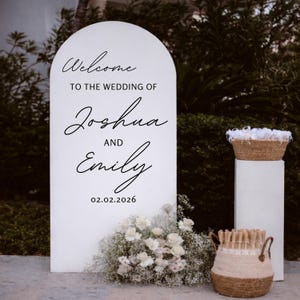 May include: A white wedding welcome sign with an arched top, featuring the text "Welcome to the Wedding of Joshua and Emily" and the date 02.02.2026. The sign is decorated with white flowers and greenery. Two woven baskets are also visible.