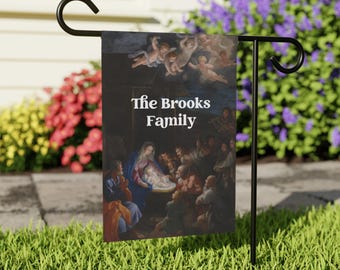 Può includere: Una bandiera da giardino con un dipinto religioso e il testo "The Brooks Family". L'opera raffigura una scena della natività con angeli, Maria, Giuseppe e altre figure. La bandiera è su un supporto in metallo nero in un giardino.