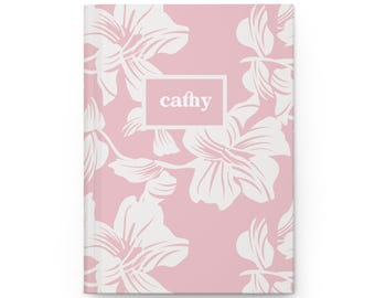 Può includere: Un diario con copertina rigida con un motivo floreale rosa e il nome "cathy" in un riquadro bianco. Il design floreale presenta grandi fiori e foglie bianche su uno sfondo rosa chiaro. Il diario è chiuso e ha un'estetica pulita e moderna.