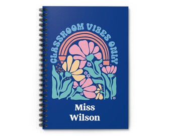 May include: A spiral-bound notebook with a navy blue cover. The cover features a floral design with a rainbow and the text "CLASSROOM VIBES ONLY". Below the design is the name "Miss Wilson" in white. The notebook is designed for teachers.