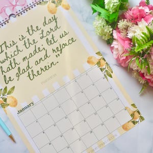 May include: A spiral-bound calendar with a light yellow background and a quote in green script. The calendar has a January page with a grid layout. Decorative elements include watercolor lemons and leaves. A light blue pen and flowers are also visible.