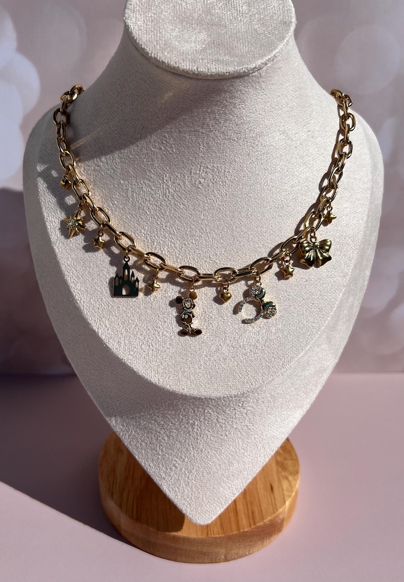 May include: Gold-toned charm necklace with various Disney-themed charms, including a castle, Mickey Mouse, and a bow. The necklace is displayed on a white bust against a soft pink background. The chain is a chunky link style.
