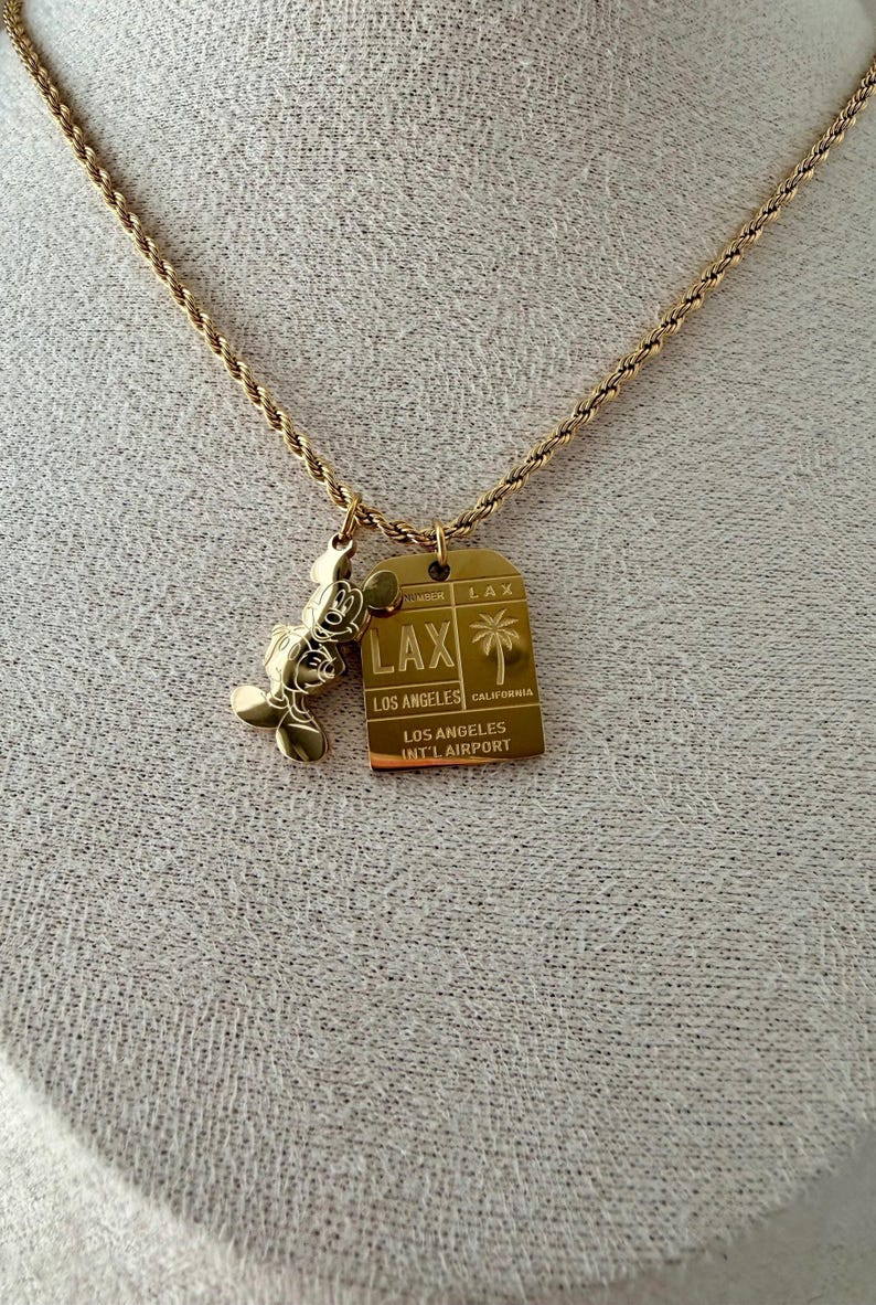 May include: A gold-toned rope chain necklace featuring two pendants: a gold-toned Mickey Mouse silhouette and a rectangular gold-toned tag engraved with LAX, Los Angeles, California, and Los Angeles Int'l Airport, including a palm tree graphic.
