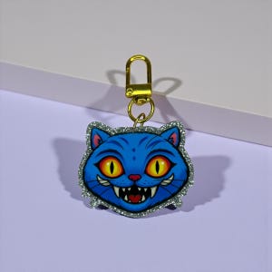 以下が含まれることがあります： 金色の留め金が付いた、青い猫の顔のキーホルダー。猫は大きな黄色の目、赤い鼻、不気味な笑顔をしています。猫の縁は銀色のグリッターで縁取られており、青との印象的なコントラストを生み出しています。
