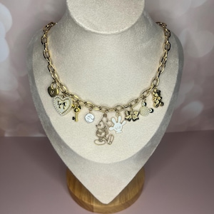 Puede incluir: Collar con colgantes dorados con varios colgantes, incluyendo un candado en forma de coraz&oacute;n, una llave, una figura de Mickey Mouse y un guante blanco. El collar se exhibe en un busto de maniqu&iacute; sobre un fondo rosa suave.