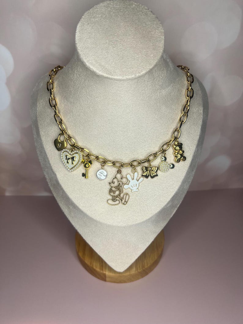 Puede incluir: Collar con colgantes dorados con varios colgantes, incluyendo un candado en forma de coraz&oacute;n, una llave, una figura de Mickey Mouse y un guante blanco. El collar se exhibe en un busto de maniqu&iacute; sobre un fondo rosa suave.