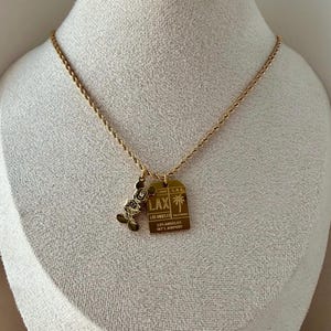 May include: A gold-toned rope chain necklace featuring two pendants: a silhouette of Mickey Mouse and a rectangular tag engraved with LAX, Los Angeles International Airport, and a palm tree graphic. The metal jewelry has a polished finish.