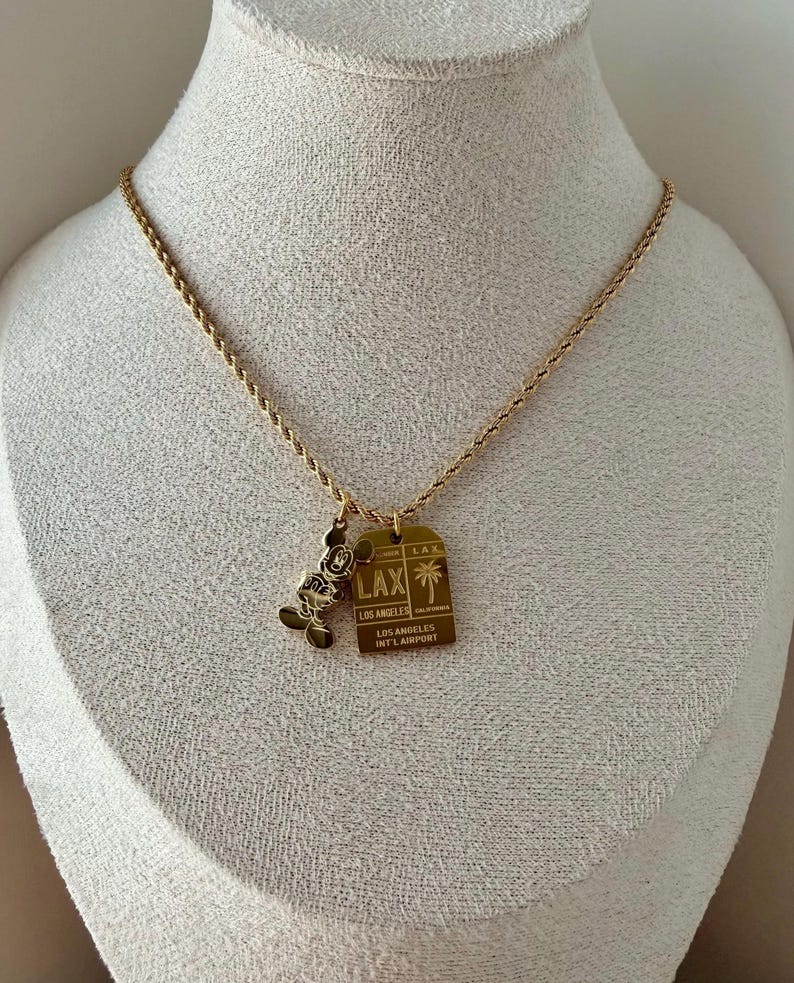 May include: A gold-toned rope chain necklace featuring two pendants: a silhouette of Mickey Mouse and a rectangular tag engraved with LAX, Los Angeles International Airport, and a palm tree graphic. The metal jewelry has a polished finish.
