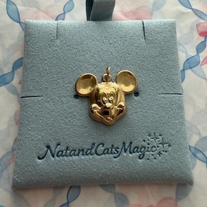 May include: A gold-toned metal pendant shaped like the head of Mickey Mouse, featuring a polished finish and a small top loop. The pendant is attached to a light blue, square-shaped felt display card with the text NatandCatsMagic printed in black.